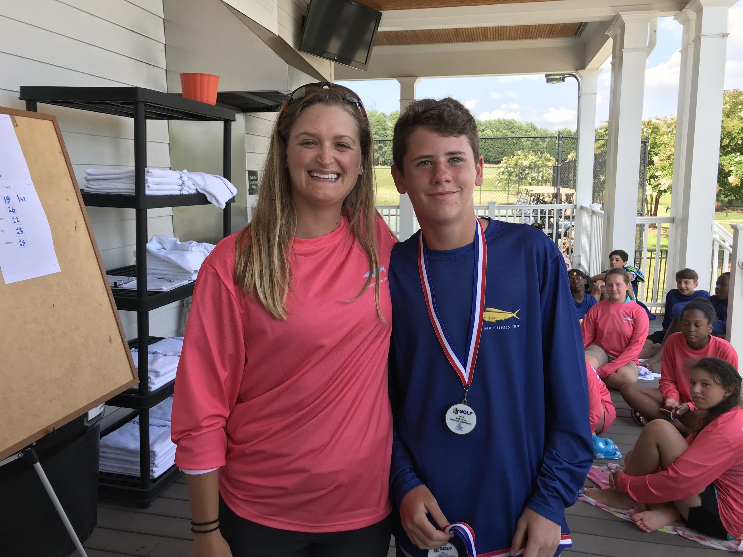 A boy wearing a medal stands beside a smiling woman on a clubhouse porch.