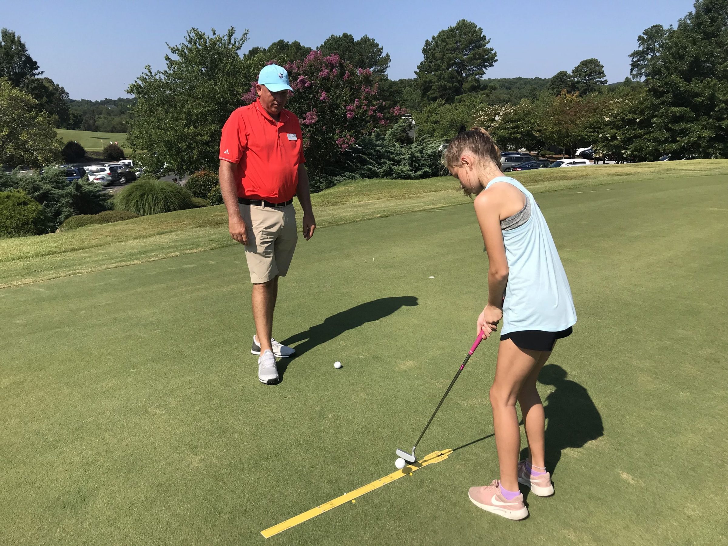 A golf instructor watches a girl putt along an alignment stick on a practice green.